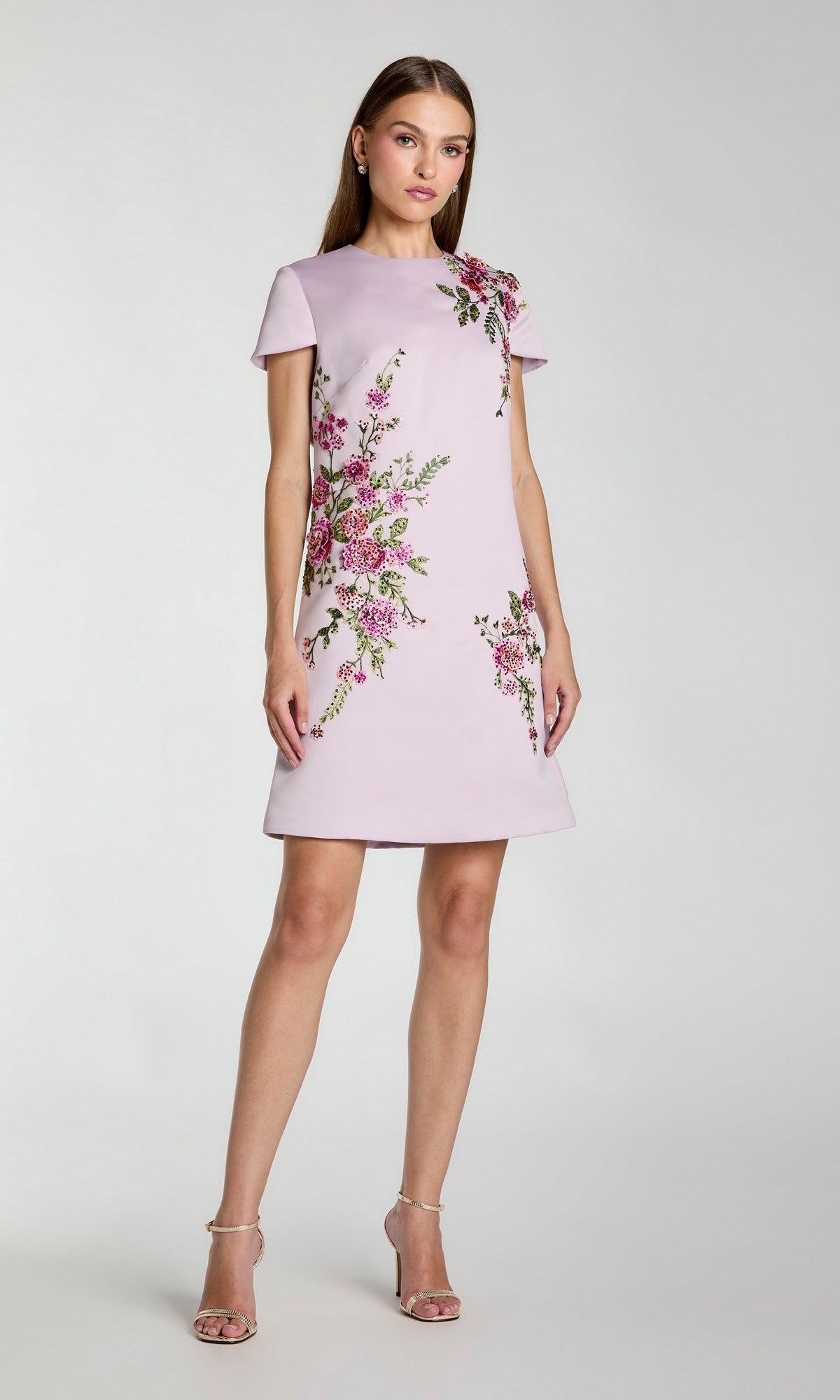 A woman wearing a short-sleeve, lilac Mac Duggal 21338 dress with floral embroidery stands against a plain light background and looks at the camera.